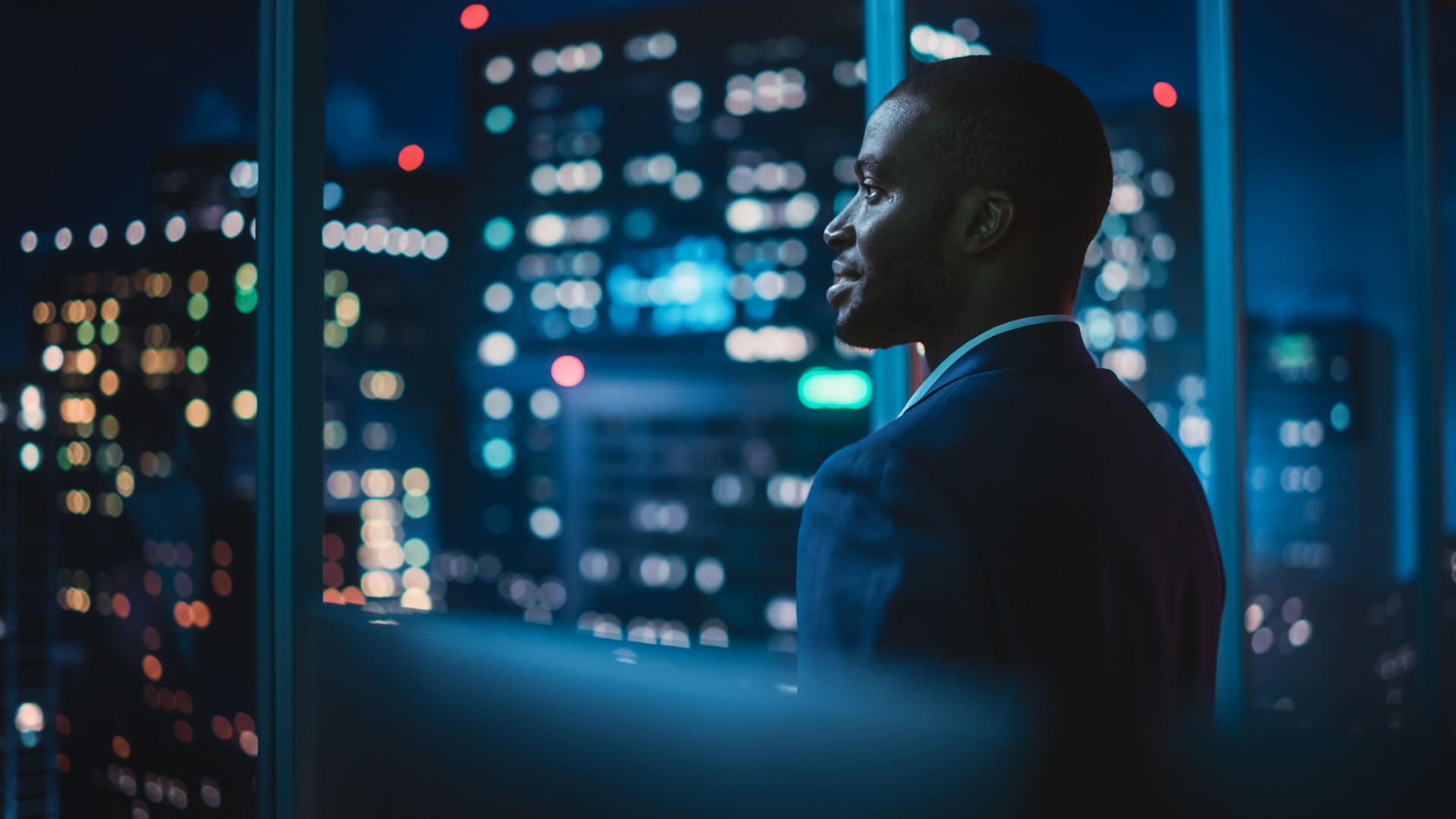 Successful Businessman Looking Out of the Window on Late Evening. Modern Hedge Fund Investor Enjoying Successful Life. Urban View with Down Town Street with Skyscrapers at Night with Neon Lights.