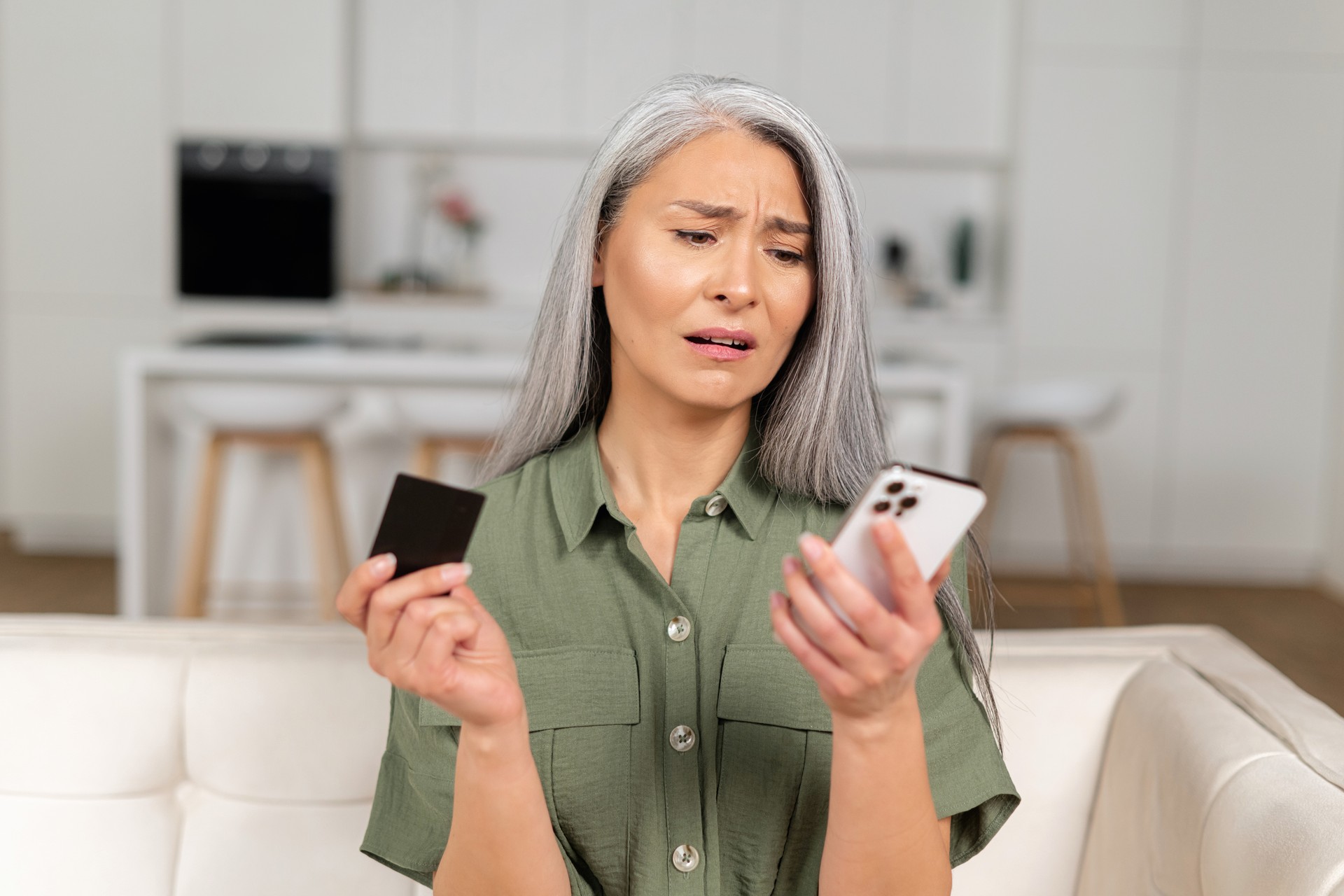 Frustrated mature woman sitting on sofa, holding smartphone and credit card